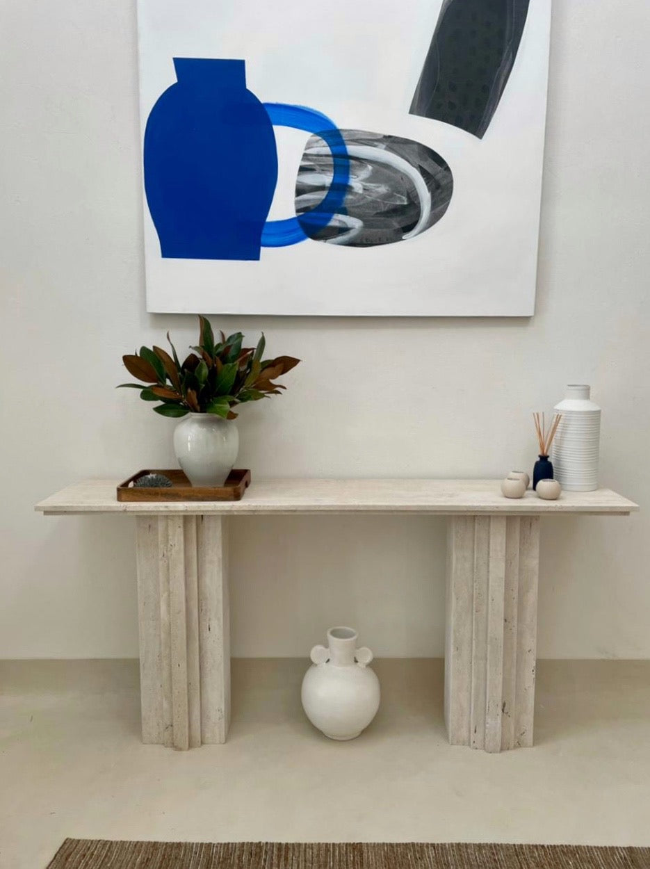 Light beige travertine console table with stepped base and top in hallway with decor and art.