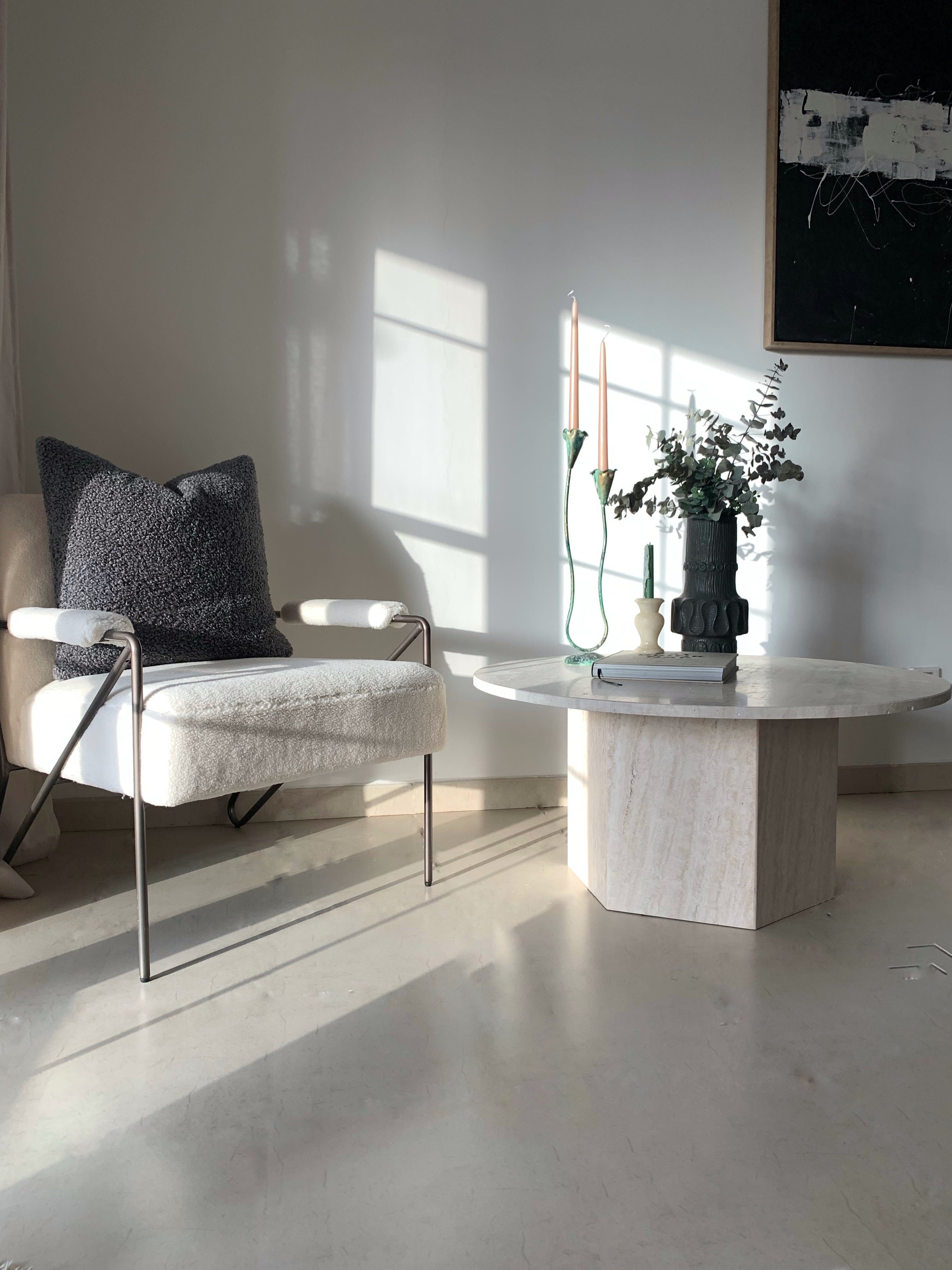 Round light beige travertine coffee table with hexagonal base next to a boucle chair.