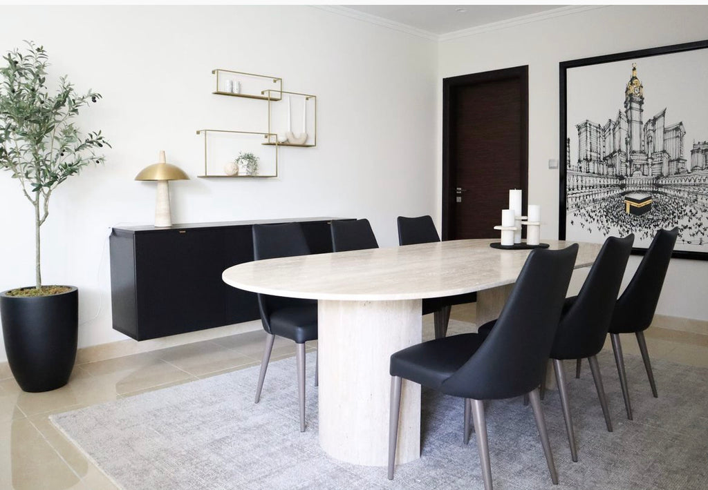 8 seater Pill shaped travertine dining table with 2 D half moon shape bases. Displayed in dining room with black leather chairs.