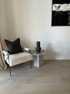 Small silver travertine side table next to comfy boucle chair and artwork.